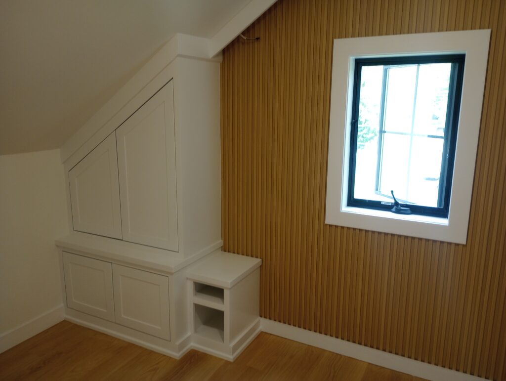A freshly painted white custom built-in cabinet flush against a sloped attic wall, paired with a vertical wood slat accent wall framing a window with crisp white trim, completed by Perfection Plus Painting in Glen Rock, New Jersey.