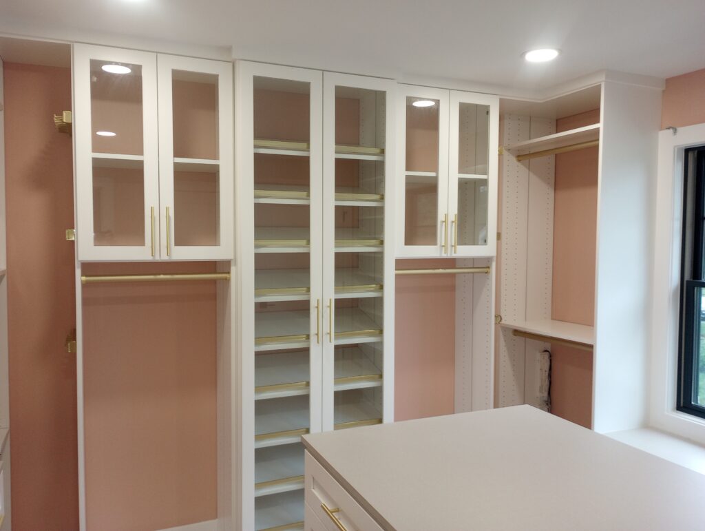A luxury custom walk-in closet featuring pristine white painted built-in cabinets, glass-front wardrobe doors, adjustable shoe shelving with gold accents, and a large center island offset by warm pink accent walls, finished by Perfection Plus Painting in Glen Rock, New Jersey.