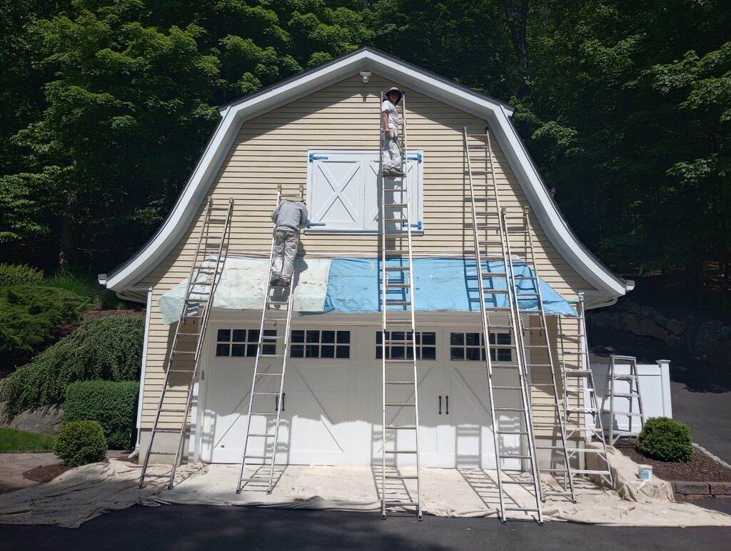 Professional house painters from Perfection Plus Painting utilizing heavy-duty extension ladders, protective scaffolding tarps, and extensive drop cloths to safely prep and paint the upper siding of a high gambrel-roof garage barn in Mahwah, New Jersey.